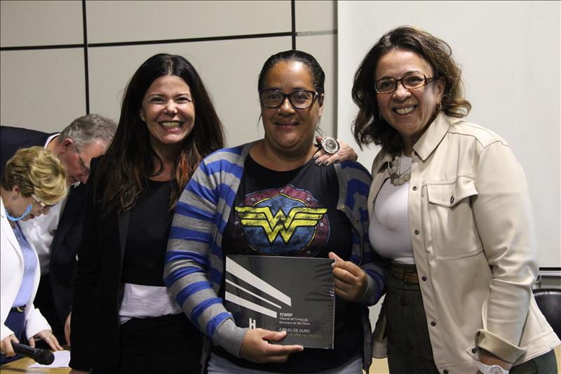 Assessora Sandra Caruso e chefe de gabinete Angélica Fernandes entregando o livro do TCMSP à munícipe